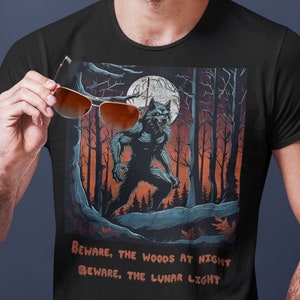 May include: Black t-shirt featuring a werewolf illustration in a forest setting with a full moon. The shirt has the text "Beware, the woods at night, beware, the lunar light." The person in the image is holding brown sunglasses.