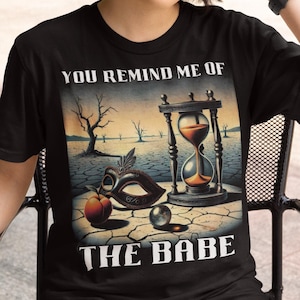 Pode incluir: Camiseta preta com uma estampa gráfica de uma pintura surrealista que apresenta uma ampulheta, uma máscara, um pêssego e uma pérola. O texto "You Remind Me Of The Babe" está impresso na camiseta.