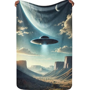 May include: A person holding a fleece blanket with a retro-style illustration of a flying saucer over a desert landscape with a planet in the background.