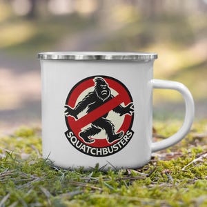Puede incluir: Taza de esmalte blanco con borde y asa plateados. La taza presenta un gráfico de una figura negra de Bigfoot dentro de un círculo rojo con una barra y el texto "Squatchbusters" debajo.