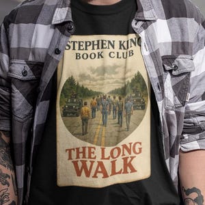 以下が含まれることがあります： 「STEPHEN KING BOOK CLUB」の文字と「THE LONG WALK」の表紙画像がプリントされた黒いTシャツ。黒いビーニー帽とグレーと白のチェック柄のシャツを着ている男性。