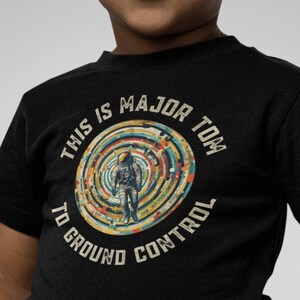 May include: Black t-shirt featuring the text "THIS IS MAJOR TOM TO GROUND CONTROL" around a circular graphic of an astronaut. The graphic has a retro, colorful design. The shirt is worn by a child.