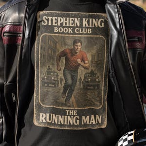 Può includere: Una persona indossa una giacca di pelle nera sopra una maglietta nera con la scritta "Stephen King Book Club" e "The Running Man". La persona tiene in mano un casco da moto a scacchi bianco e nero. Jeans blu completano il look.