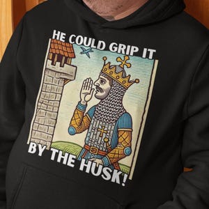 May include: A black hooded sweatshirt featuring a graphic print of a medieval king wearing a crown and chainmail, standing near a stone tower. The text above and below the illustration reads, HE COULD GRIP IT BY THE HUSK! The hoodie has long sleeves.