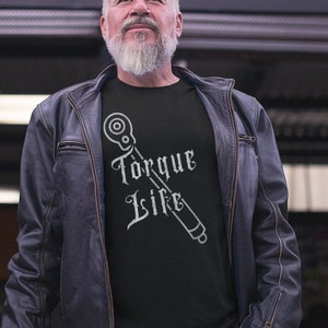 May include: A black t-shirt with the words "Torque Life" and a wrench graphic in white. The person is wearing a black leather jacket. The background includes a shop with motorcycles.