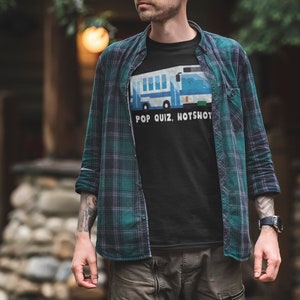以下が含まれることがあります： 青と白のバスのグラフィックと「POP QUIZ, HOTSHOT」のテキストが入った黒いTシャツ。シャツは、緑と青のチェック柄のシャツの下に着ています。その人は腕時計とカーゴパンツを着用しています。