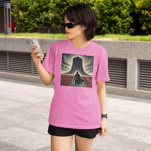 May include: A woman wearing a pink t-shirt with a graphic of a person walking towards a large, dark structure in a field of red flowers. The graphic is surrounded by a white border.
