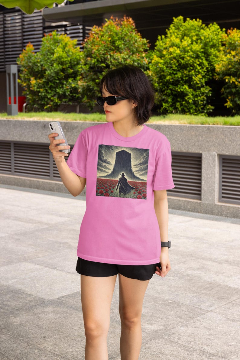 May include: A woman wearing a pink t-shirt with a graphic of a person walking towards a large, dark structure in a field of red flowers. The graphic is surrounded by a white border.