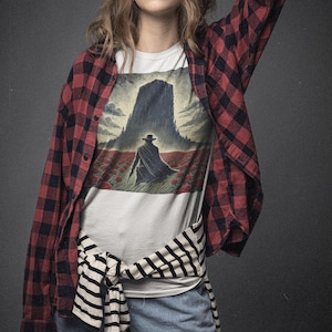 May include: A woman wearing a white t-shirt with a graphic print of a man walking towards a tall building in a field of red flowers. She is wearing a red and black plaid shirt tied around her waist and blue jeans.