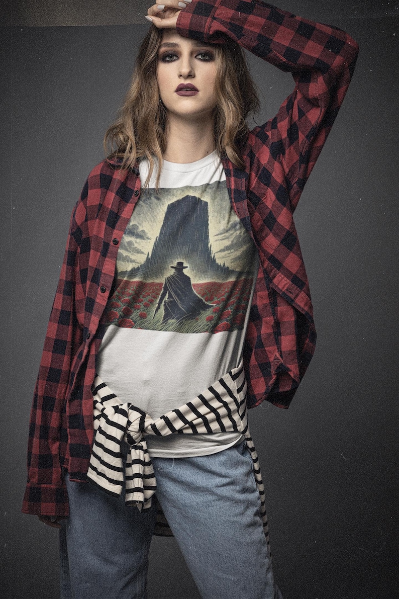 May include: A woman wearing a white t-shirt with a graphic print of a man walking towards a tall building in a field of red flowers. She is wearing a red and black plaid shirt tied around her waist and blue jeans.