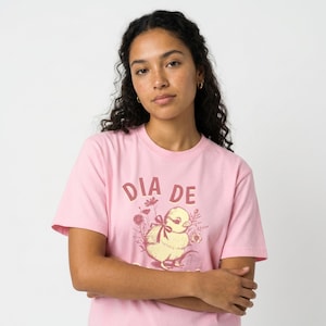 Op de afbeelding: Een lichtroze t-shirt met de tekst "DIA DE" boven een afbeelding van een gele kuiken met een strik en bloemen. Het shirt is gemaakt van een zacht materiaal en heeft korte mouwen.