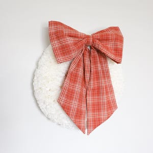 May include: A white, fluffy wreath with a large, red and white plaid bow. The bow is centered at the top of the wreath and has long, flowing tails. The wreath is hanging on a white wall.