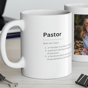 May include: White ceramic mug with a black and white definition of the word "Pastor" and a photo of two young girls hugging. The mug has a black handle.
