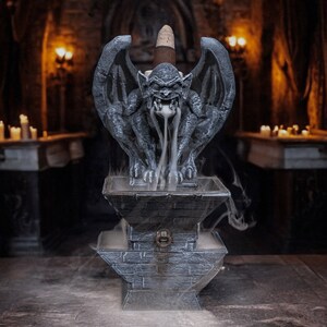 May include: A gray stone gargoyle incense burner with a cone incense stick burning inside. The gargoyle is sitting on a stone base with a small opening in the front. Smoke is coming out of the gargoyle's mouth and swirling around the burner.