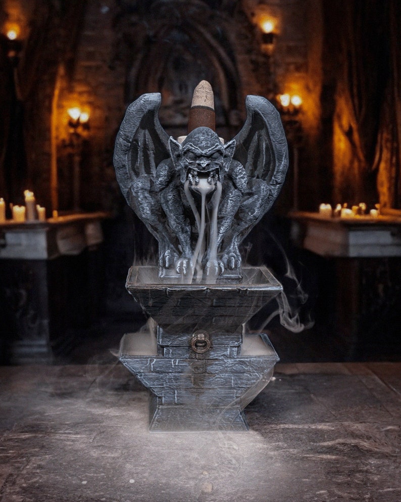 May include: A gray stone gargoyle incense burner with a cone incense stick burning inside. The gargoyle is sitting on a stone base with a small opening in the front. Smoke is coming out of the gargoyle's mouth and swirling around the burner.