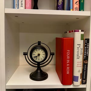 May include: A black, decorative desk clock with a circular face and Roman numerals sits on a white shelf. The clock is flanked by books, including a red book titled "FOUR IN A ROW" and a book by Jodi Picoult.