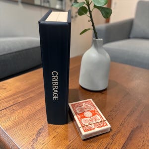 May include: A navy blue cribbage board with the word "CRIBBAGE" in white lettering. A deck of playing cards is next to the board. A small white vase with a plant is in the background.