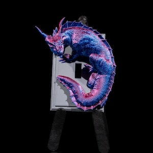 May include: A blue and pink dragon figurine perched on a white light switch cover. The dragon is curled up with its tail wrapped around its body. The light switch cover is mounted on a black easel.
