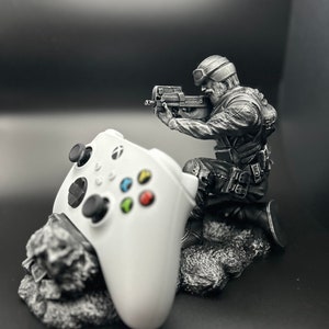 May include: A white video game controller is held by a silver figurine of a soldier holding a gun. The figurine is standing on a rocky base.