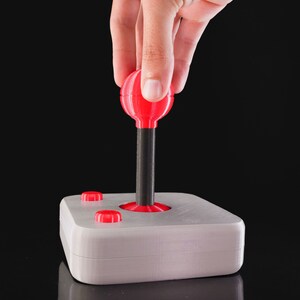 May include: A white joystick with a red ball on top and two red buttons. The joystick is on a black surface.