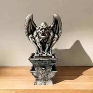 May include: A silver gargoyle statue with wings, perched atop a stone base with a ring handle. The gargoyle has a fierce expression with sharp teeth and claws.