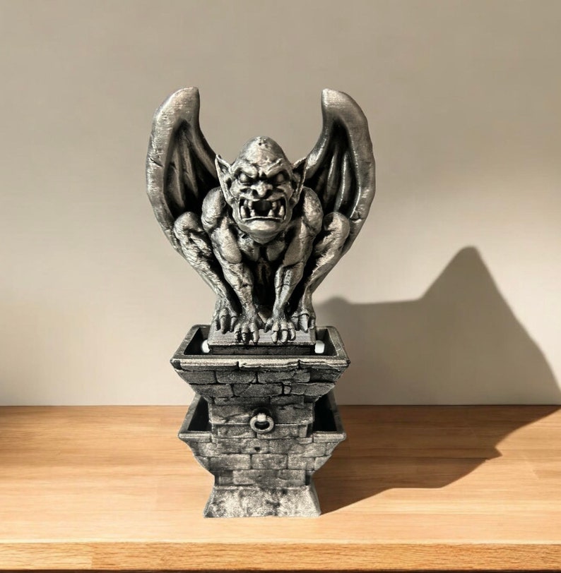 May include: A silver gargoyle statue with wings, perched atop a stone base with a ring handle. The gargoyle has a fierce expression with sharp teeth and claws.