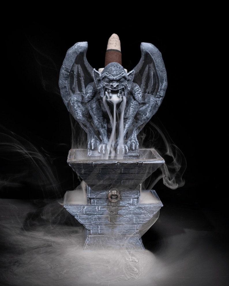May include: A gray stone gargoyle incense burner with a white incense cone burning inside. The gargoyle is sitting on a stone base with a small door on the front. Smoke is coming out of the gargoyle's mouth and swirling around the burner.