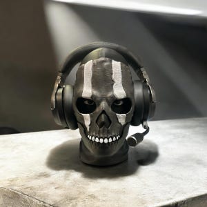 May include: A black and white skull mask with a headset on top. The mask has a menacing expression with a wide mouth and sharp teeth. The headset is black with silver accents.