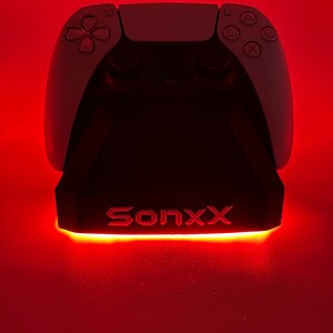 May include: A white PlayStation 5 controller sits on a black charging stand with a red light. The stand has the word "Sonxx" printed on it.
