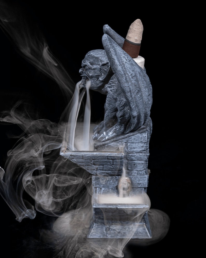 May include: A gray stone gargoyle incense burner with a cone incense stick burning. Smoke flows from the gargoyle's mouth and down the front of the burner. The burner is on a black background.