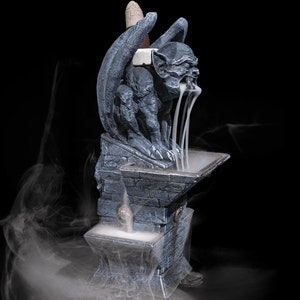 May include: A gray stone gargoyle incense burner with a winged design. The incense is burning and smoke is flowing from the gargoyle's mouth. The burner is on a stone base.