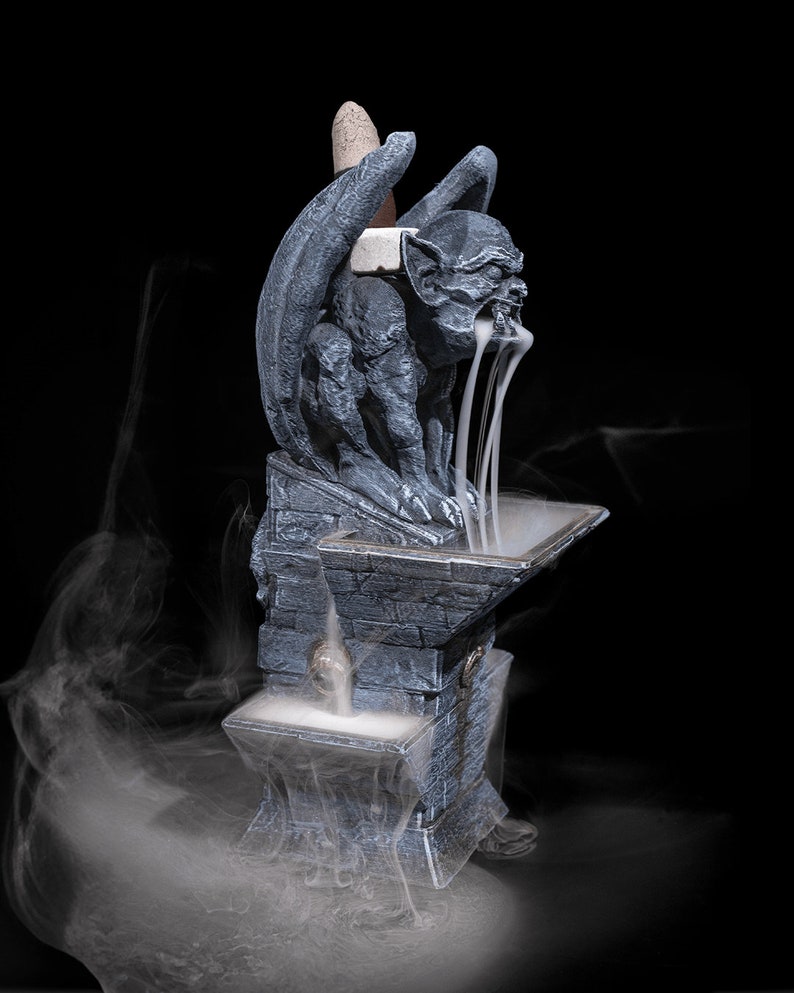 May include: A gray stone gargoyle incense burner with a winged design. The incense is burning and smoke is flowing from the gargoyle's mouth. The burner is on a stone base.