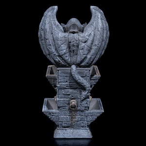 May include: A gray stone fountain with a gargoyle figure with wings at the top. The fountain has three tiers with a decorative design.
