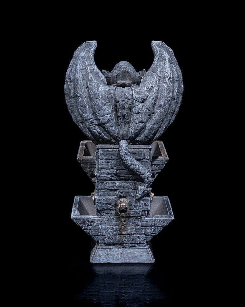 May include: A gray stone fountain with a gargoyle figure with wings at the top. The fountain has three tiers with a decorative design.