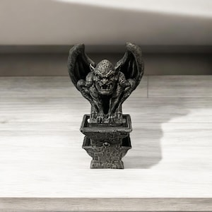 May include: A black gargoyle statue with wings perched on a stone base. The gargoyle has a menacing expression and appears to be made of a hard, durable material.