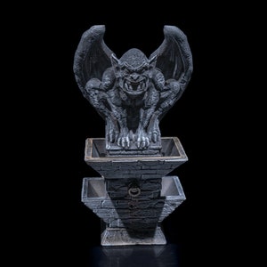 May include: A gray stone gargoyle statue with wings perched atop a gray stone fountain with two tiers. The gargoyle has a menacing expression with sharp teeth and claws.