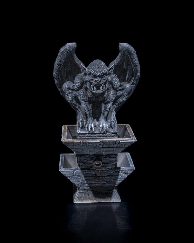 May include: A gray stone gargoyle statue with wings perched atop a gray stone fountain with two tiers. The gargoyle has a menacing expression with sharp teeth and claws.