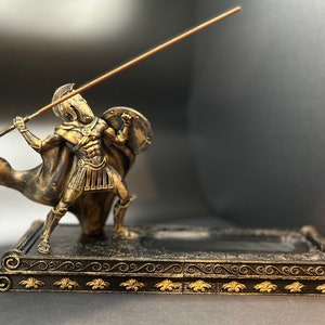May include: A gold-colored statue of a warrior holding a spear and shield, standing on a black and gold decorative base. The statue is a decorative incense holder.