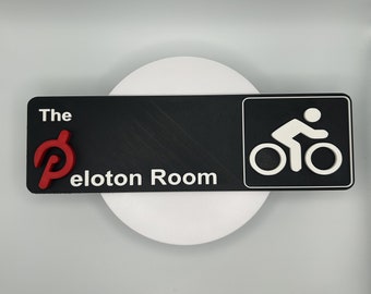 May include: Black sign with white text that reads "The Peloton Room" and a red Peloton logo. A white silhouette of a person riding a bicycle is on the right side of the sign.
