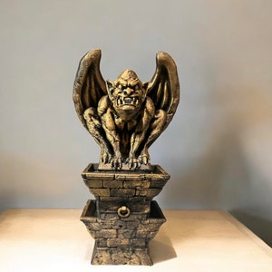 May include: A gold gargoyle statue with wings perched atop a two-tiered stone base. The gargoyle has a menacing expression and is sculpted in a realistic style.
