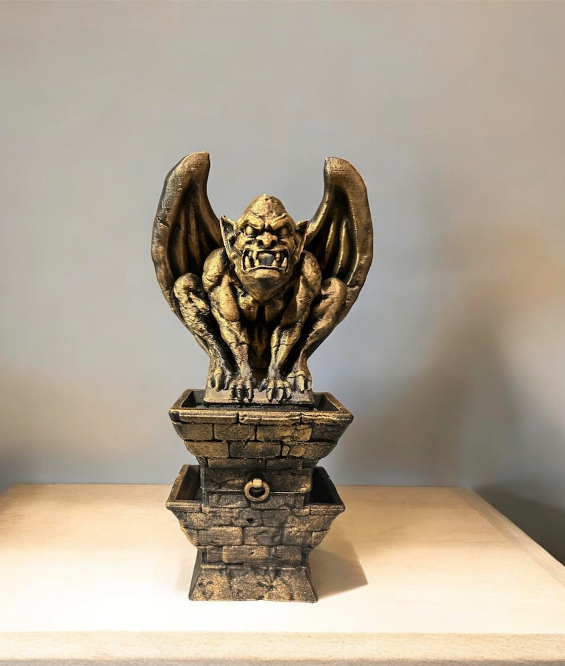 May include: A gold gargoyle statue with wings perched atop a two-tiered stone base. The gargoyle has a menacing expression and is sculpted in a realistic style.