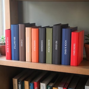 May include: A selection of board games, such as "Four in a Row" and "Checkers," are displayed on a wooden shelf. The game boxes are in various colours, with white text labels. The games are in a row.