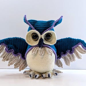May include: A crocheted owl figurine with a dark teal head and body, cream-coloured belly, and large black eyes. The wings are spread wide, featuring dark teal tops, purple trim, and light grey feathered edges. It has a dark beak and feet.