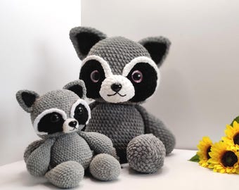 May include: Two handmade crochet raccoon plush toys in varying sizes, featuring grey yarn bodies, white muzzle accents, black eye patches, and dark plastic eyes. The larger raccoon sits upright while the smaller one is positioned in front.