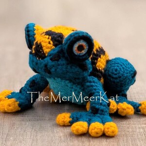 May include: A crocheted frog toy featuring a vibrant pattern of deep blue and bright yellow yarn. The frog has large, round eyes with orange and black irises. The text TheMerMeerKat is printed in white across the center of the frog's body.
