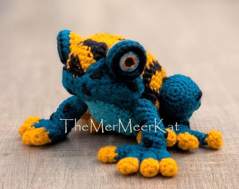 May include: A crocheted frog toy featuring a vibrant pattern of deep blue and bright yellow yarn. The frog has large, round eyes with orange and black irises. The text TheMerMeerKat is printed in white across the center of the frog's body.