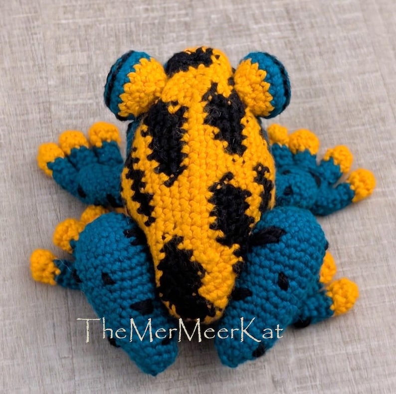 May include: A crocheted frog toy featuring a bright yellow back with black spots, blue limbs, and yellow toes. The text TheMerMerKat is embroidered in white at the bottom. The stuffed animal has a textured yarn surface and a rounded shape.