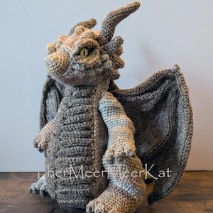 Könnte beinhalten: Eine gehäkelte Drachenfigur in Braun- und Grautönen mit texturierten Schuppen, großen Flügeln und gelben Augen. Das Wesen hat Hörner auf dem Kopf und der Text "TherMeerMeerKat" ist in weißer Schrift im unteren Bereich zu sehen.