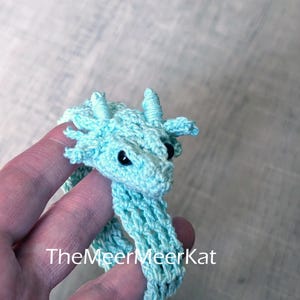 May include: A light blue crocheted dragon bracelet featuring a detailed head with small horns, side fins, and black bead eyes. The flexible, open-weave band wraps around the wrist. The text TheMeerMeerKat is printed on the image.