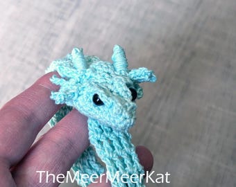 May include: A light blue crocheted dragon bracelet featuring a detailed head with small horns, side fins, and black bead eyes. The flexible, open-weave band wraps around the wrist. The text TheMeerMeerKat is printed on the image.